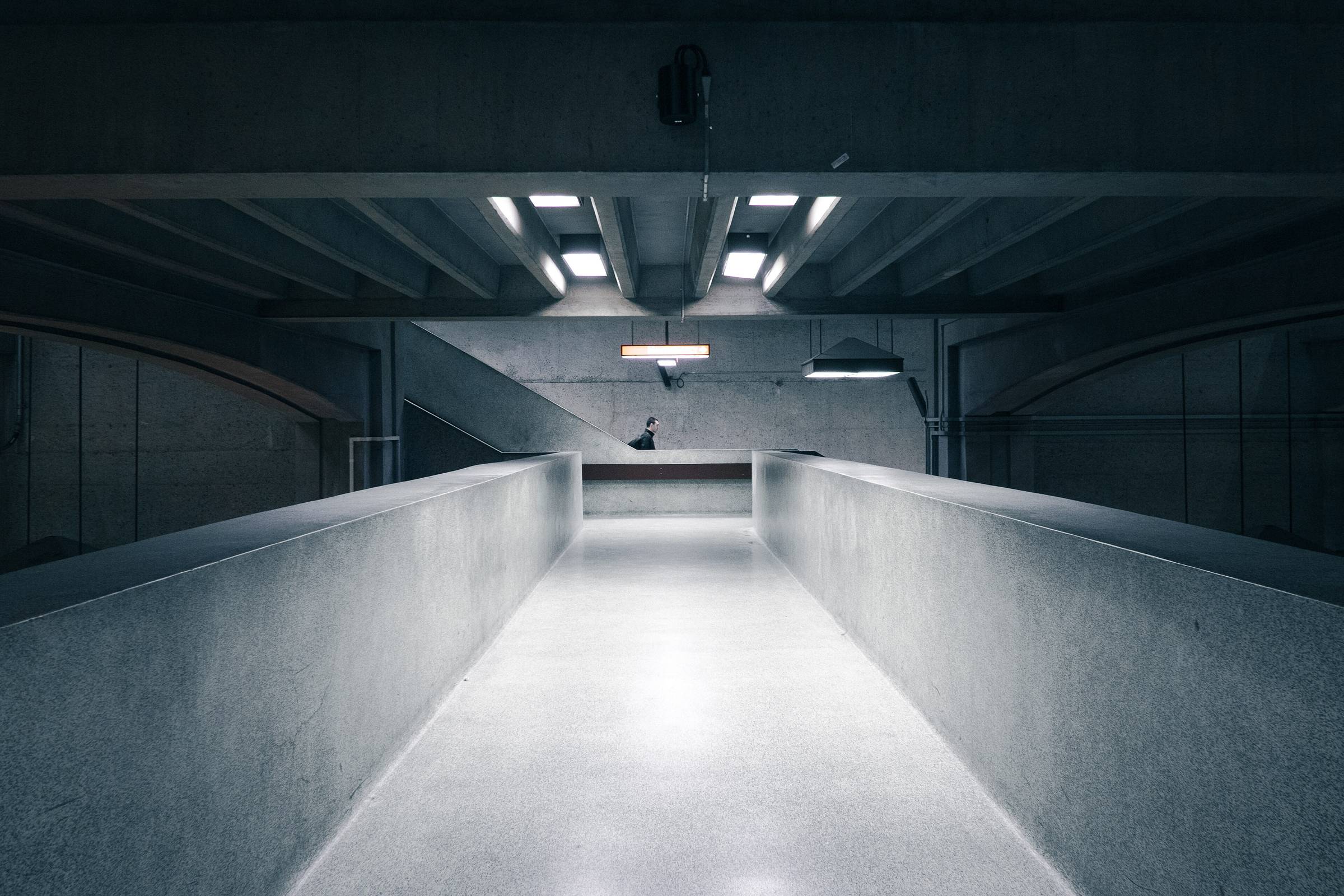 A figure walking a long concrete corridor lit from the ceiling, photographed in monochrome.