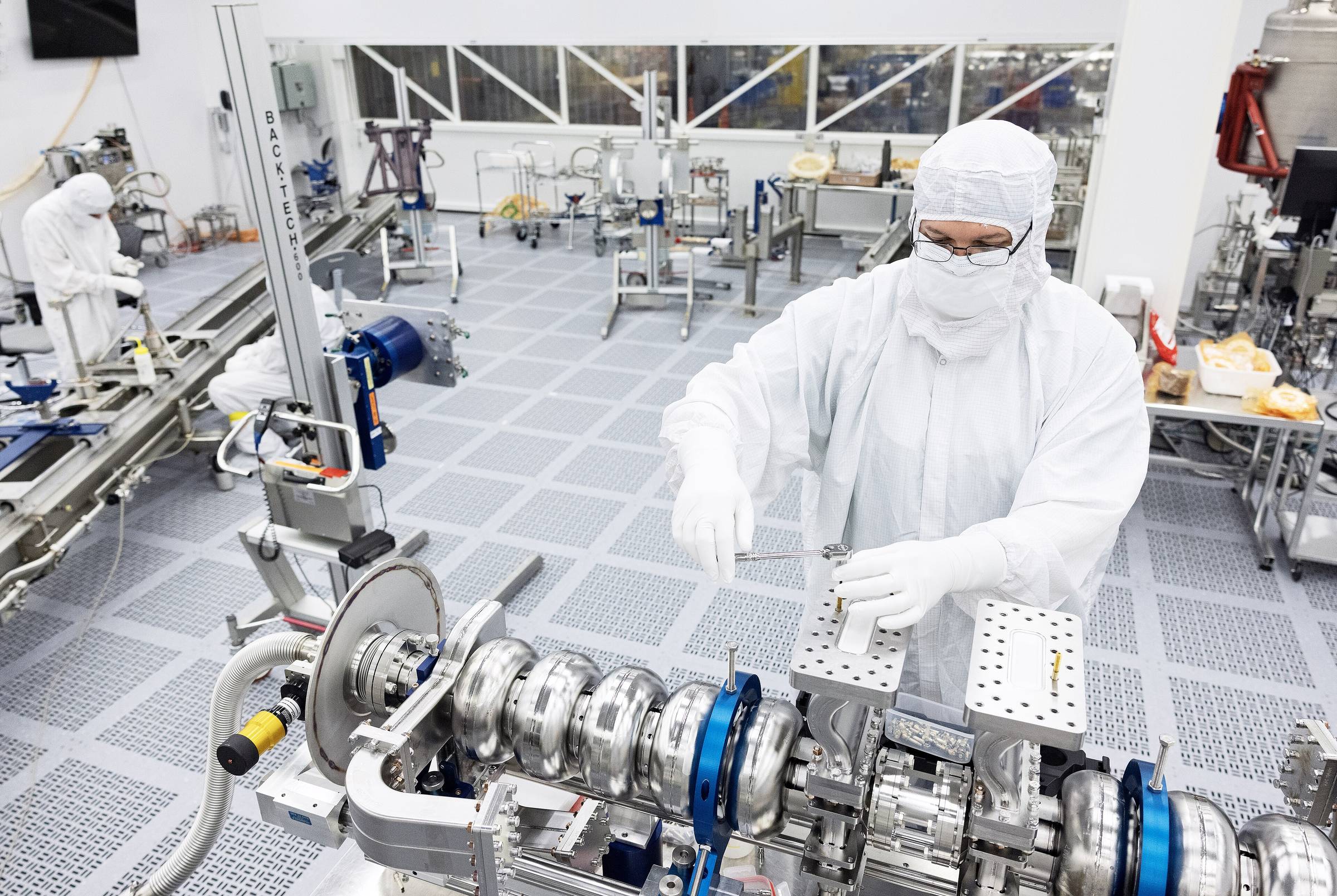 Technicians in full clean-room gowning working at a bank of instruments.