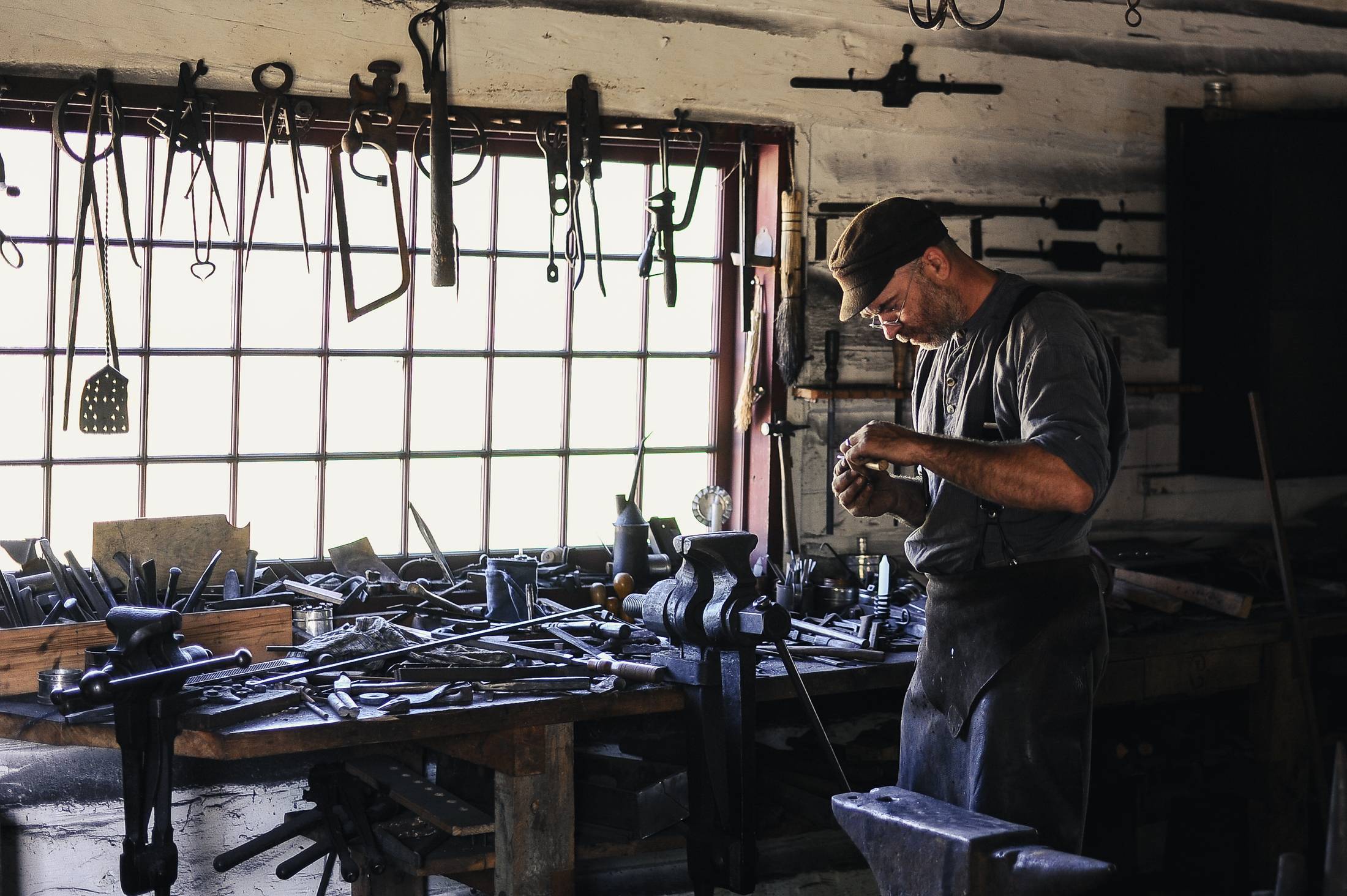 A man alone at a workbench in a small shop, rows of tools hung on the wall behind him.