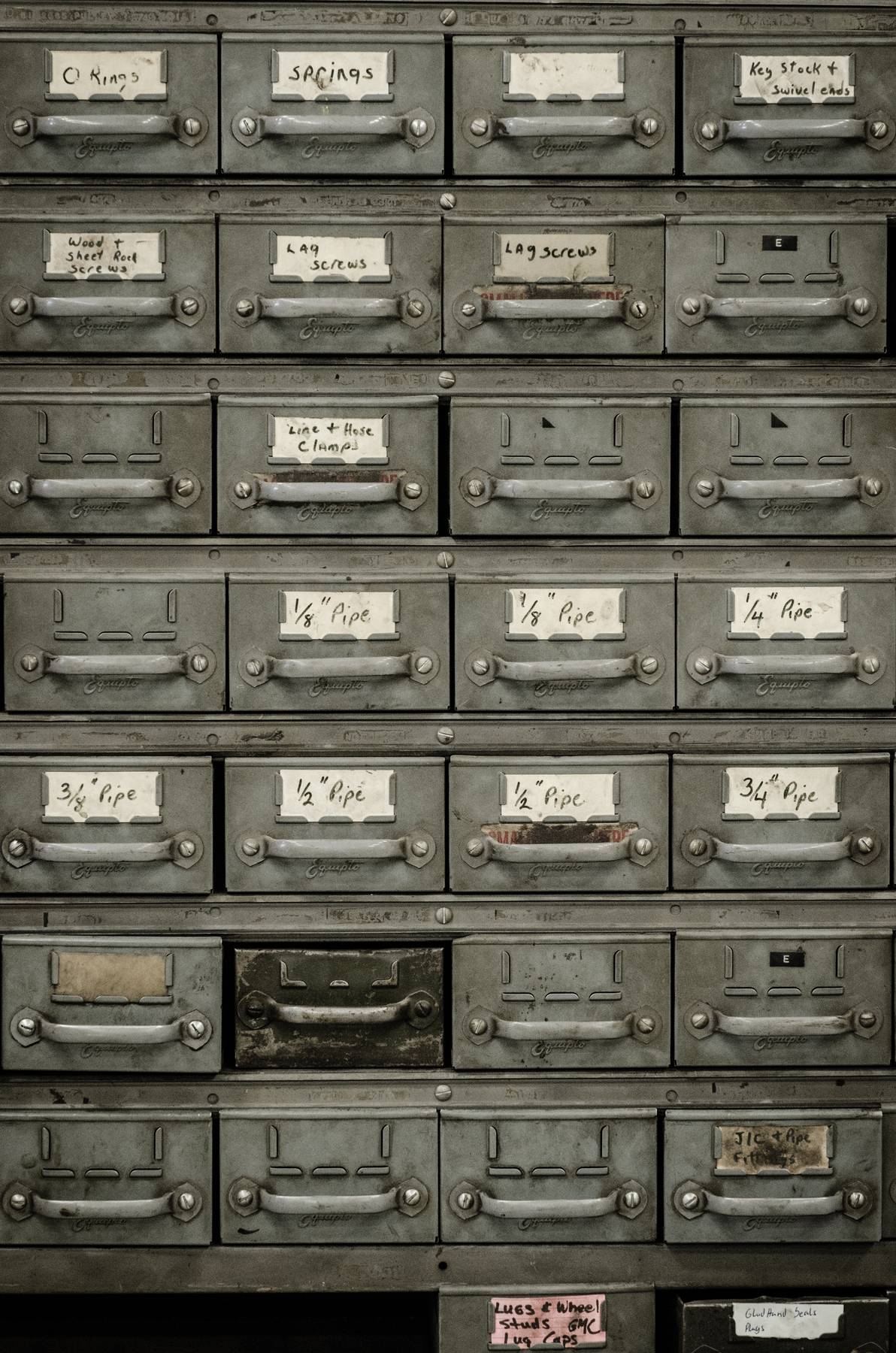 A wall of small labelled parts drawers in an old supply room, handles worn bright.
