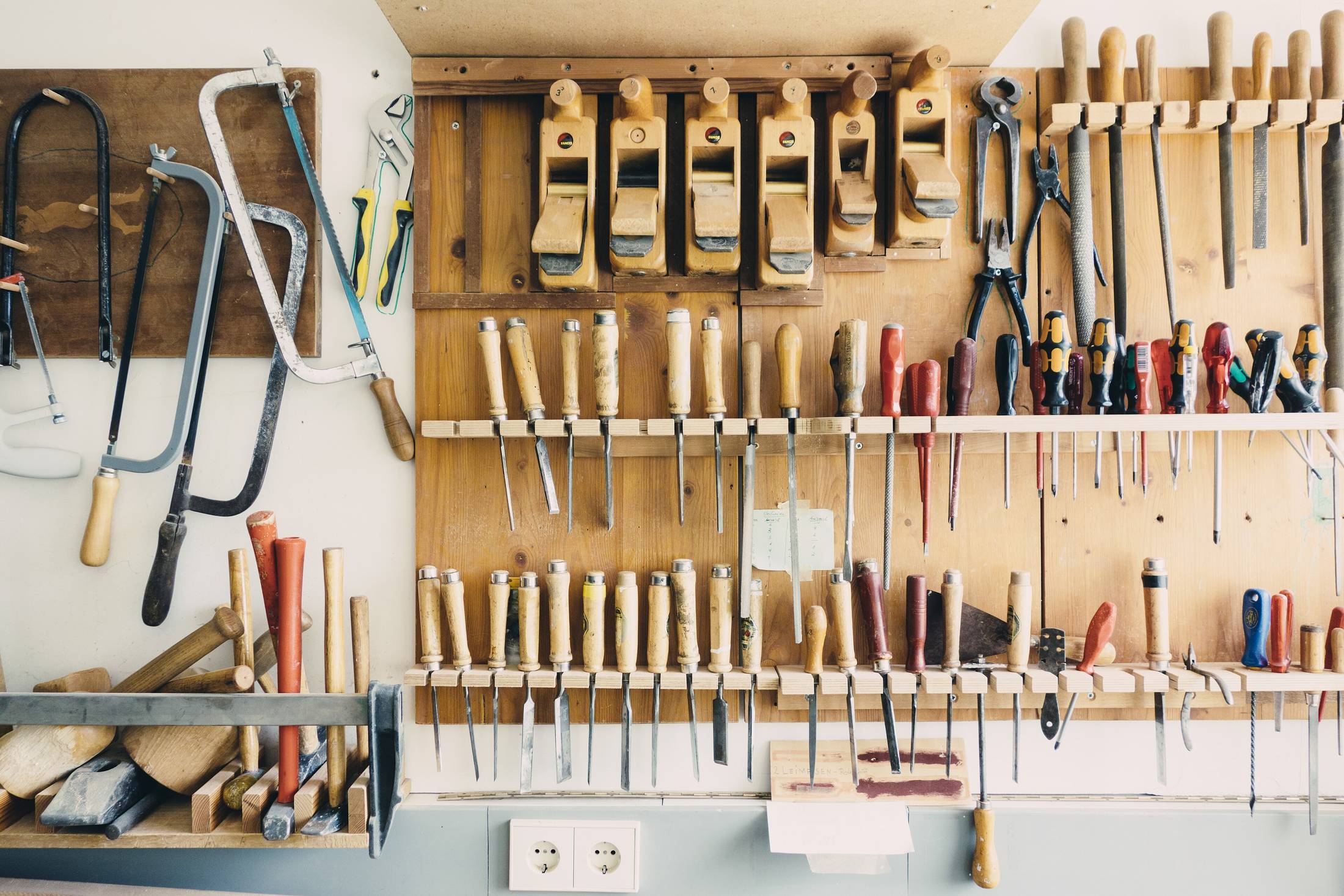 A workshop tool wall: saws, chisels, pliers and files hung in order on a painted board.